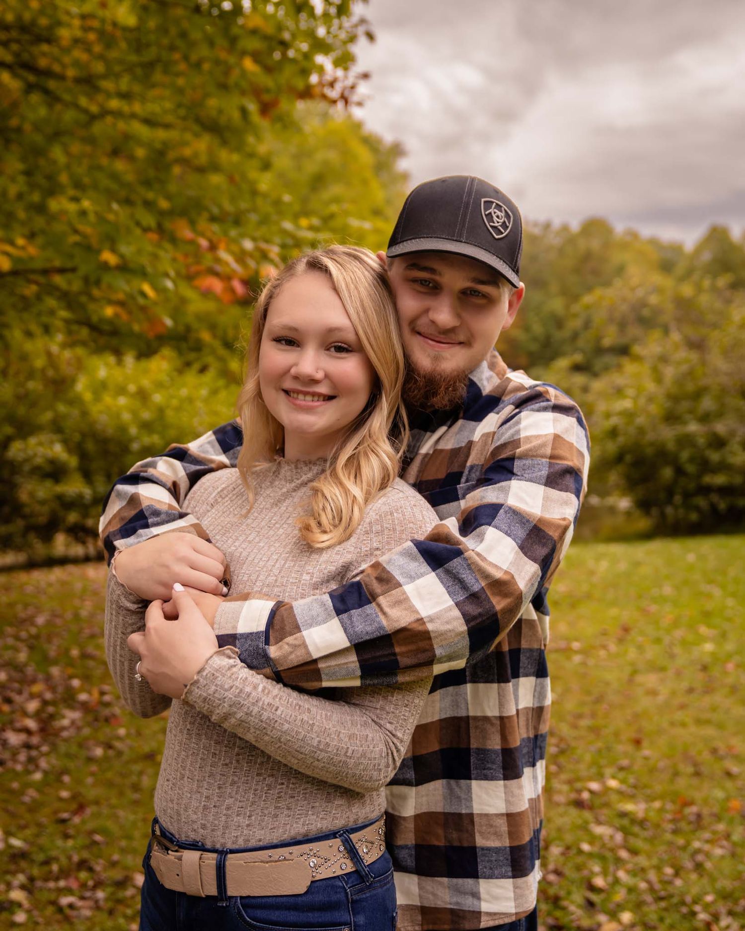 A young couple embraces warmly in a fall park setting, with the man hugging the woman from behind.