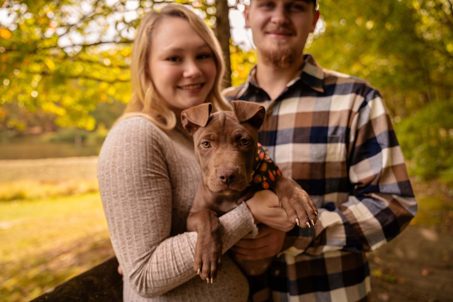 A young blonde woman holds a small brown puppy while a man stands beside her in a colorful fall outdoor setting.