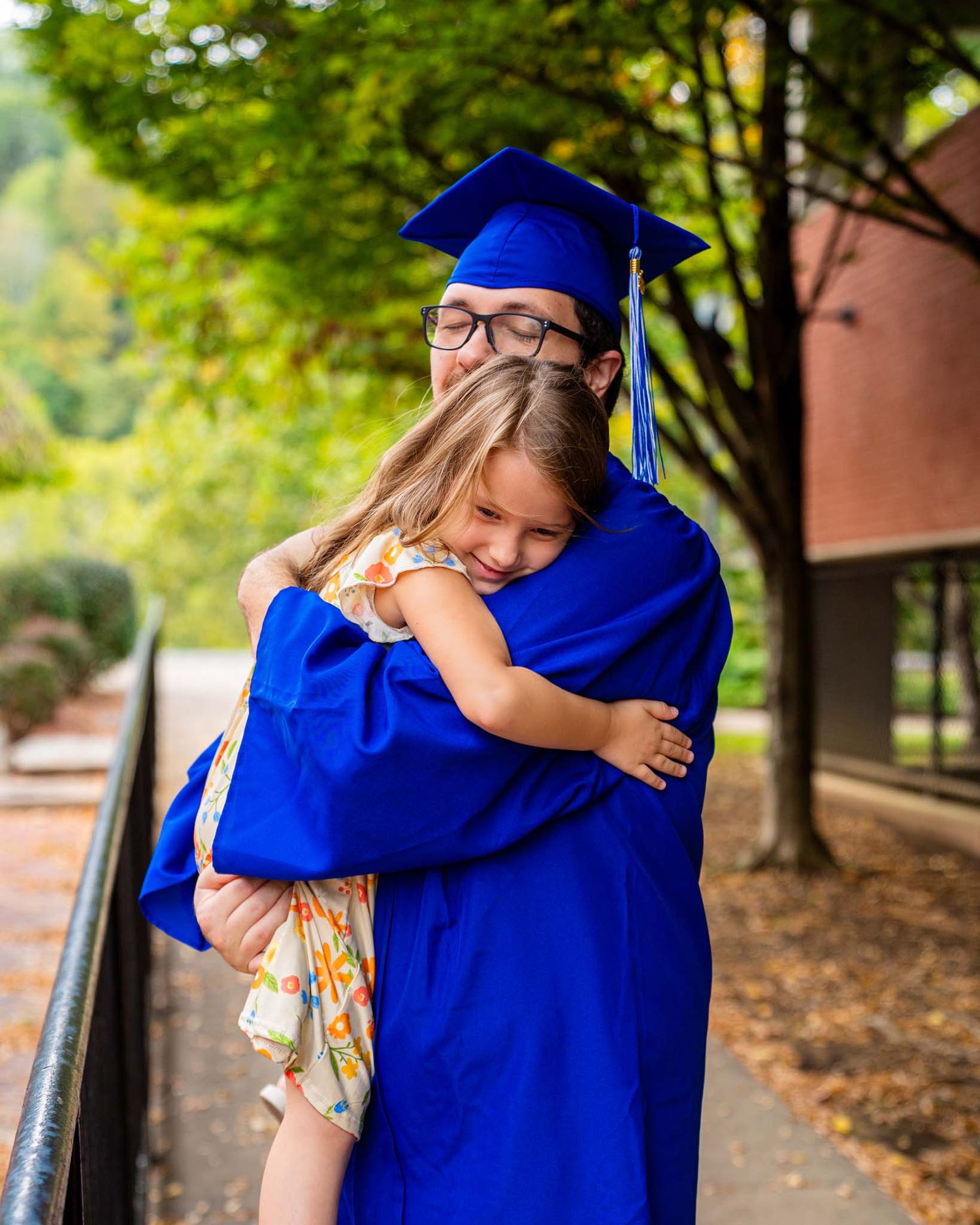 Male graduate in blue cap and gown hugging young girl on walkway.