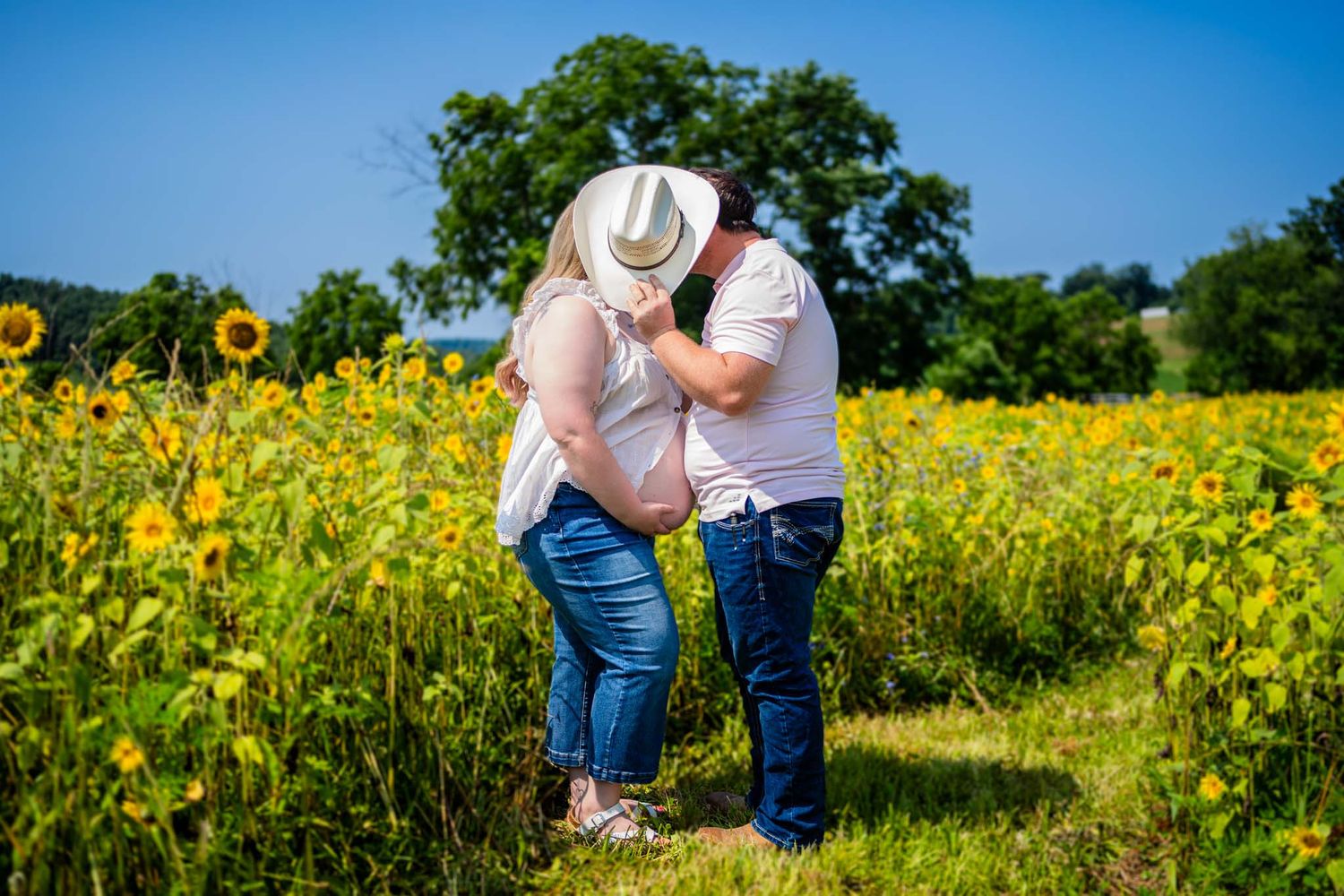 Couple kissing in a sunflower field wearing white hats and jeans on a sunny day.
