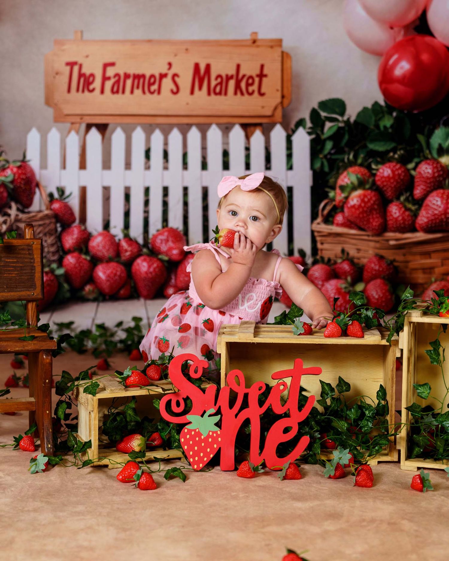 Baby girl in strawberry outfit sits in crate at Farmer's Market themed first birthday Sweet One cake smash photo.