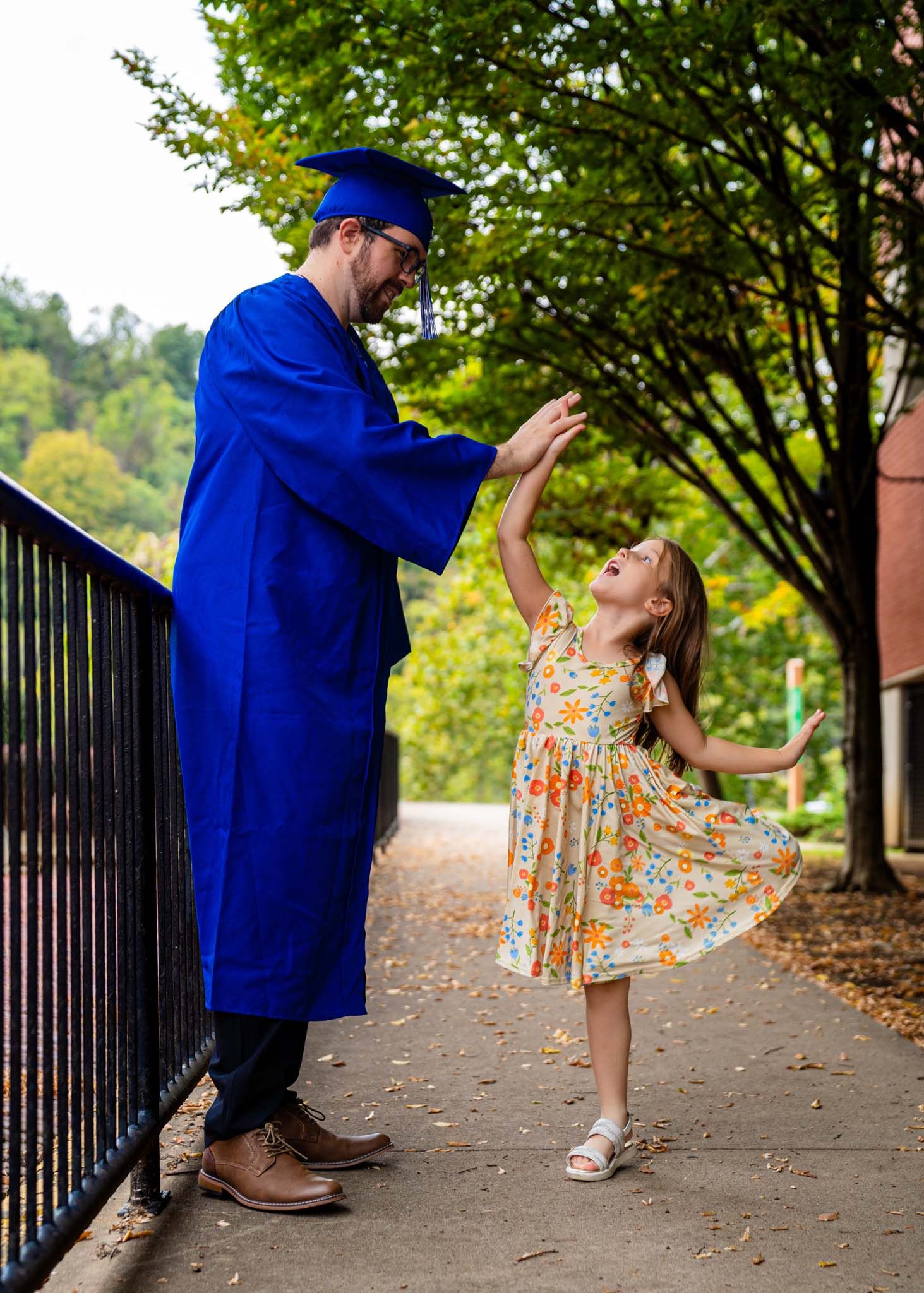 Graduate in blue cap and gown twirling a young girl in a floral dress outdoors on a tree-lined path.