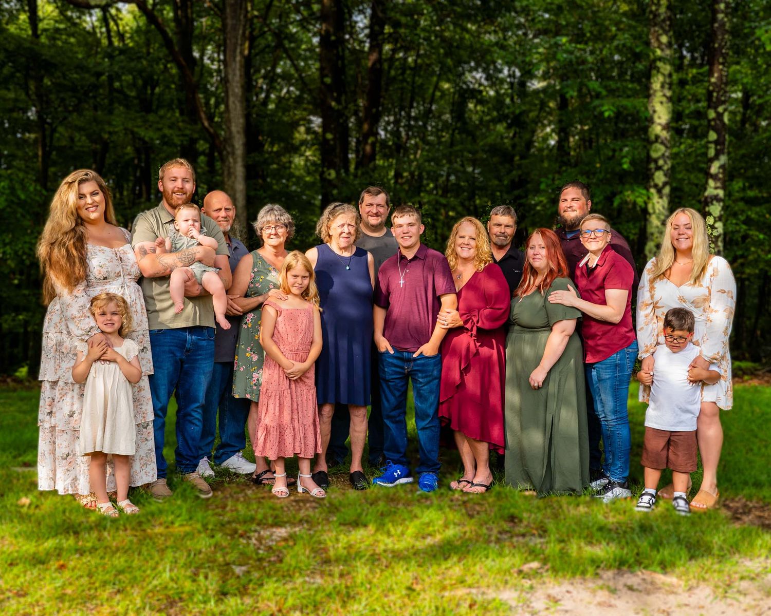 Large extended family group portrait outdoors, smiling together in coordinating outfits among lush green trees.