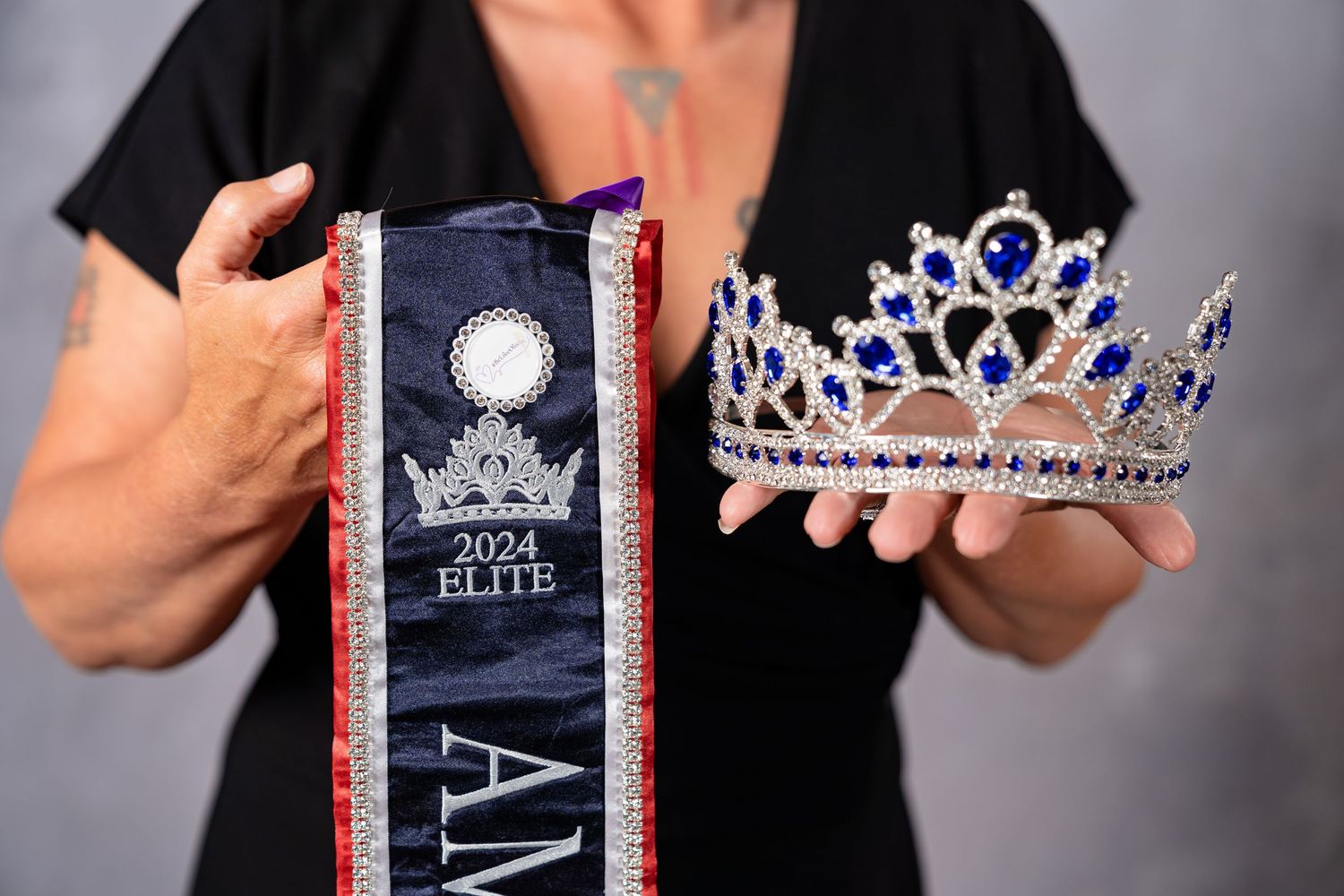 Woman holding a 2024 Elite pageant sash and jeweled crown with blue gemstones.
