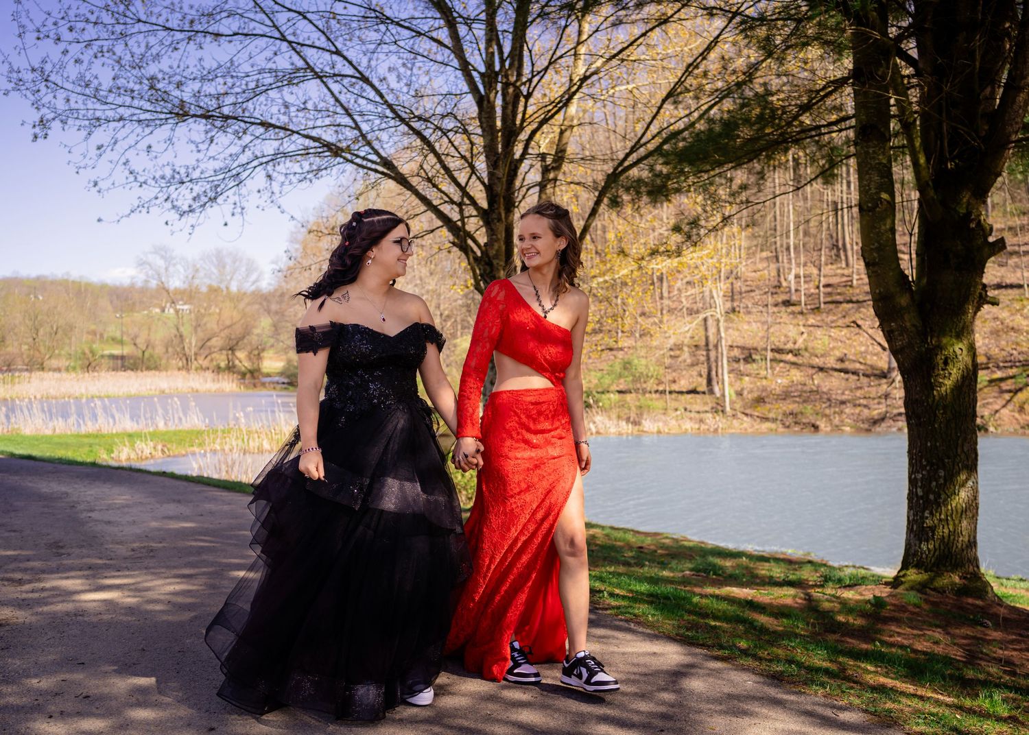 Two young women in formal gowns walking hand in hand along a lakeside path in spring.