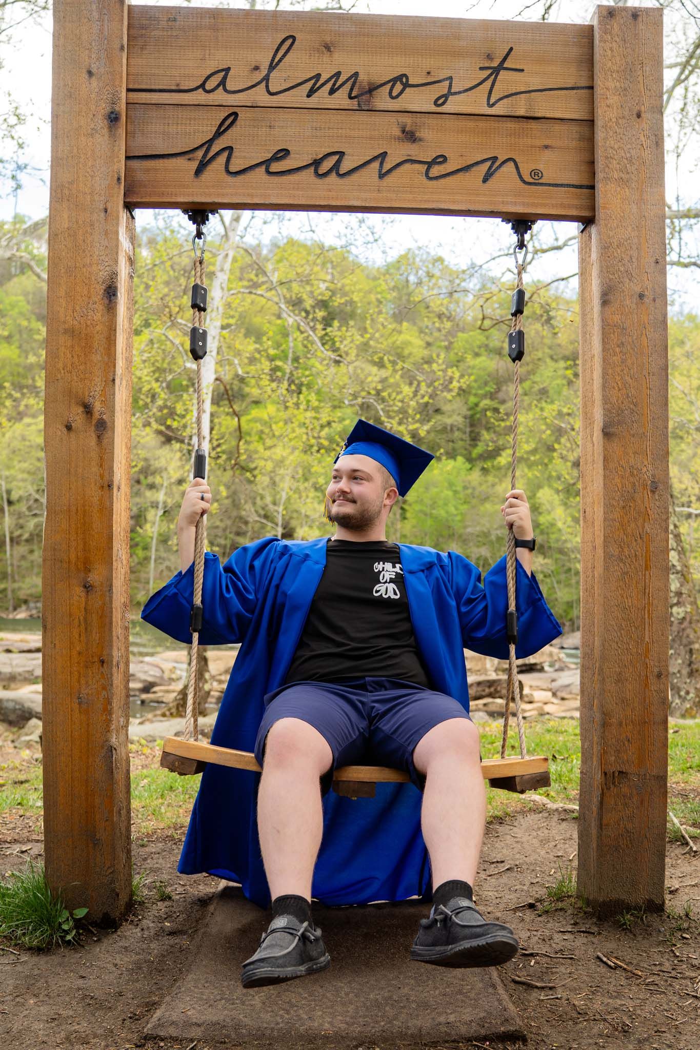 Male graduate in blue cap and gown sitting on Almost Heaven swing in outdoor setting.