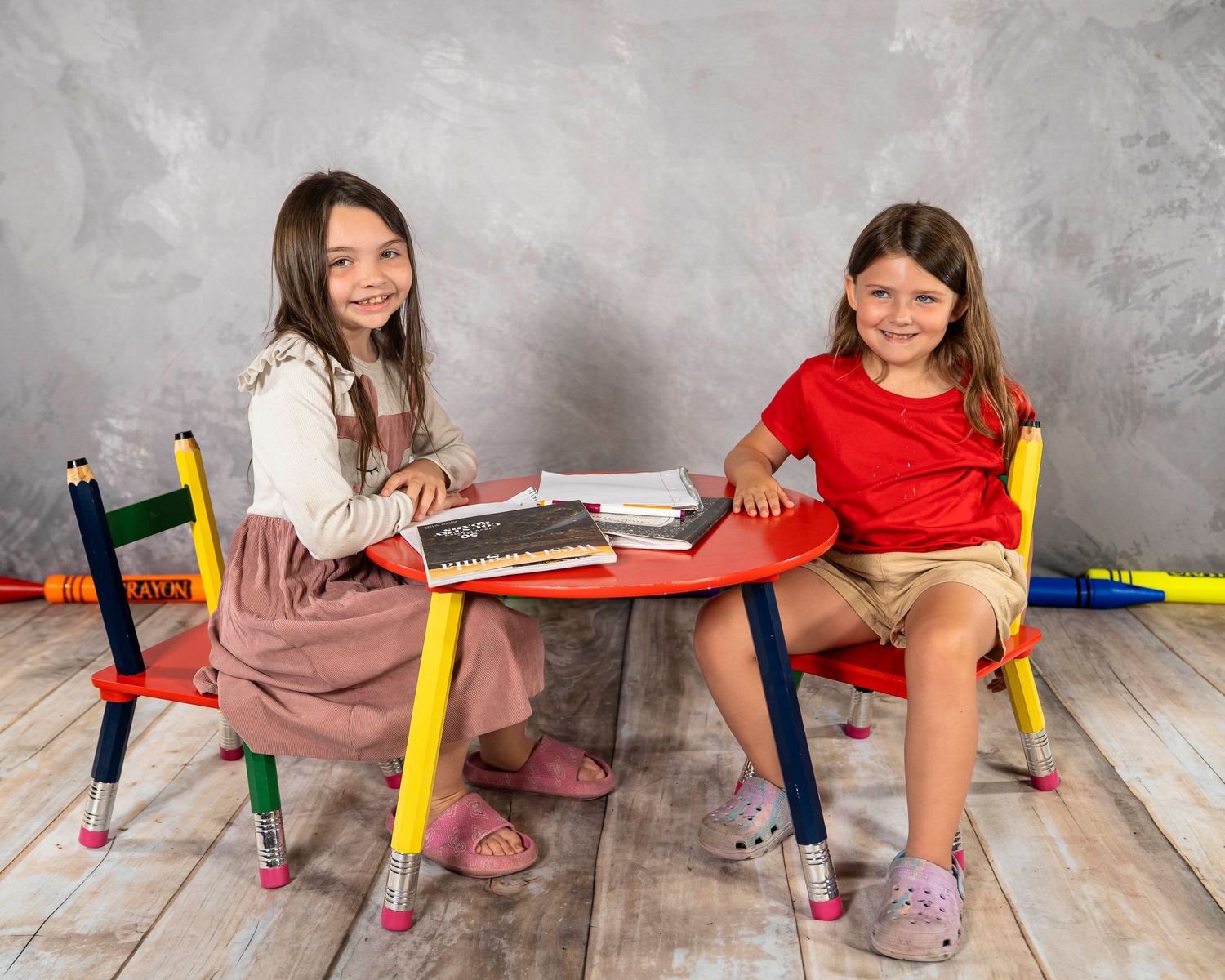 Two girls sitting at a colorful pencil-themed kids table and chair set, smiling and reading books together.