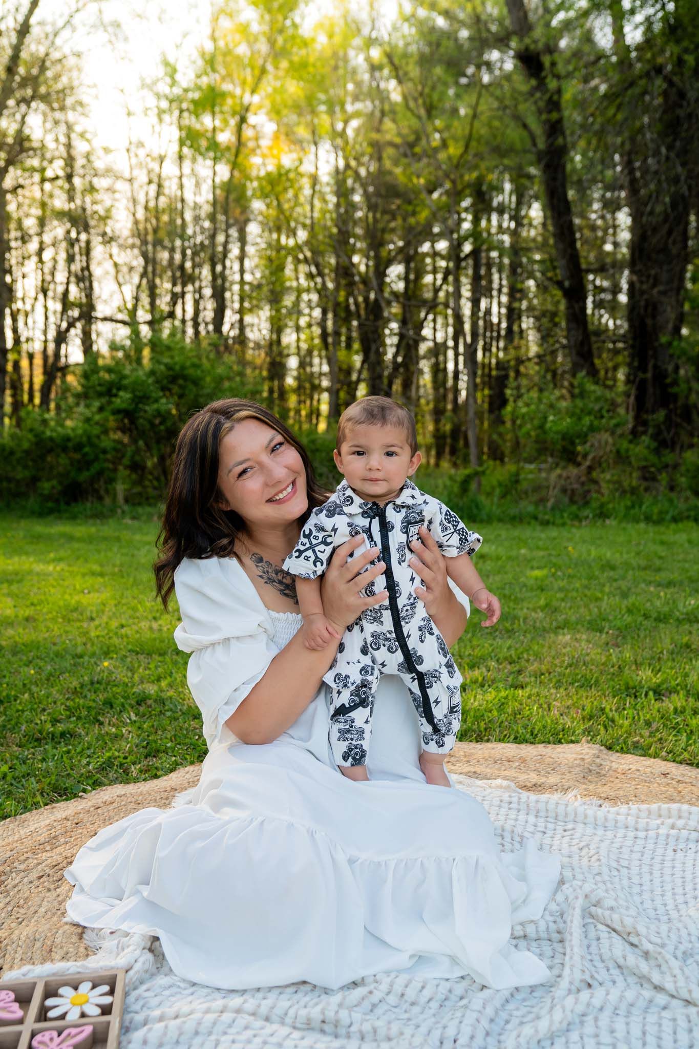 A smiling mother in a white dress holds her baby in a black and white outfit on a blanket in a sunny green park.
