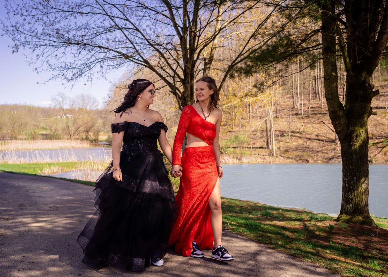 Two young women in formal gowns, one black and one red, walk along a scenic lakeside path surrounded by trees.