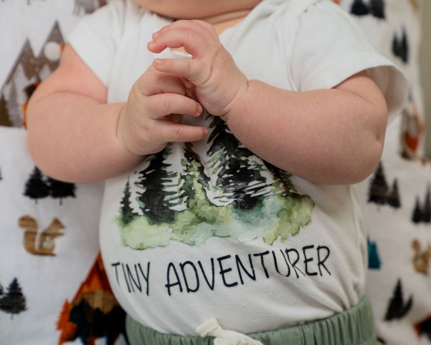 Baby wearing a white 'Tiny Adventurer' onesie with forest print, paired with green pants.