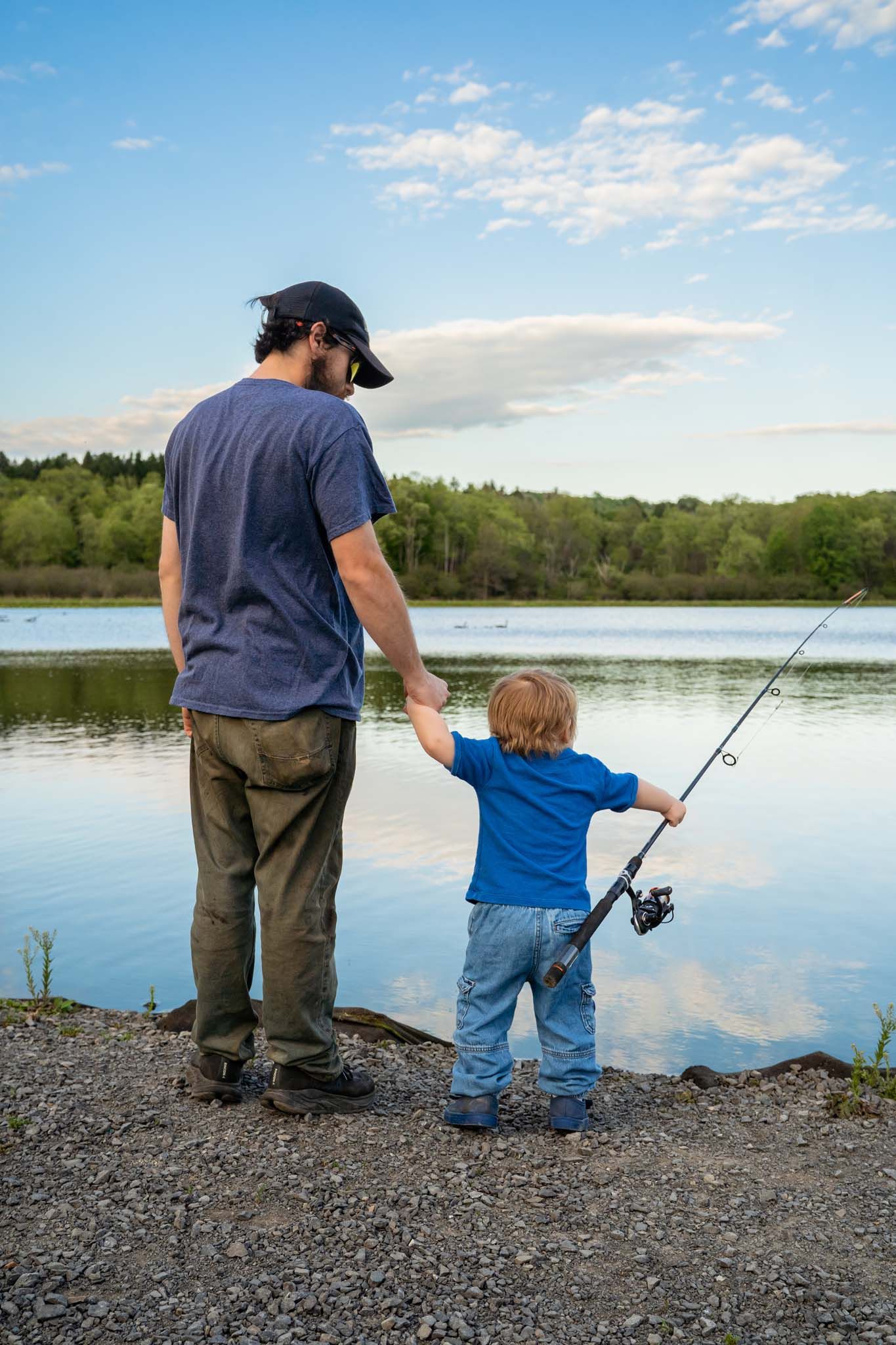 Father and young child fishing together at peaceful lake shore during golden hour with blue sky.