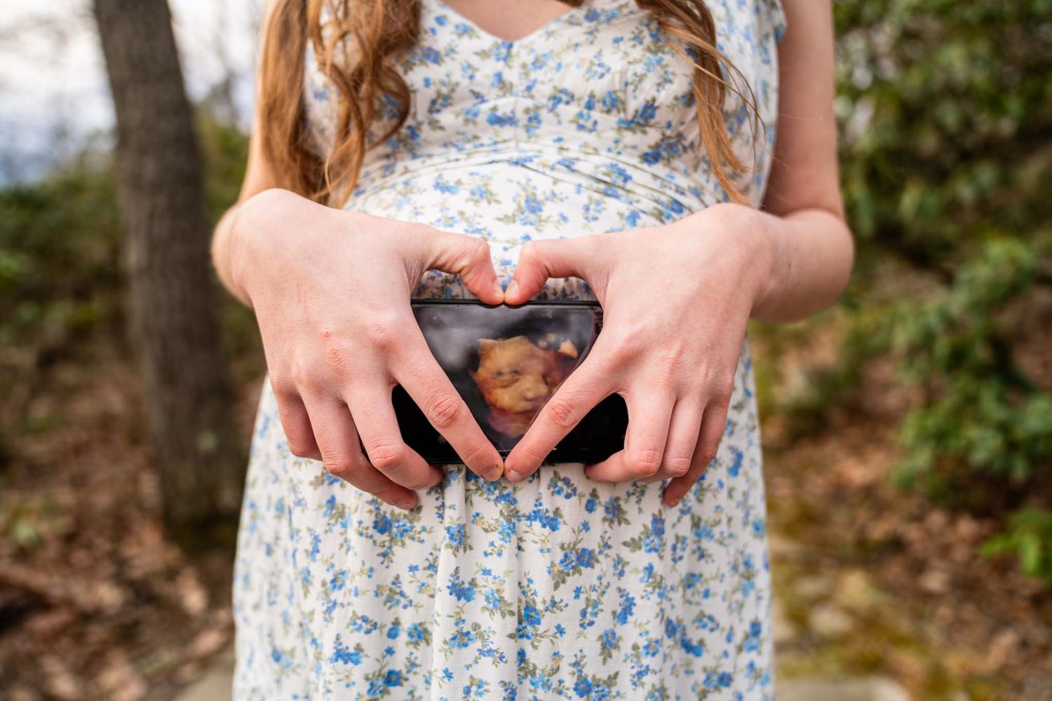 Pregnant woman forms heart shape with hands around ultrasound photo in outdoor maternity portrait.