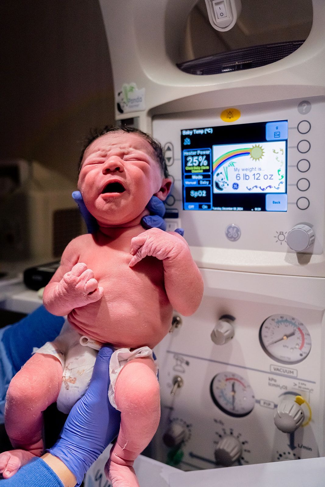 Crying newborn baby being held up after birth, weighing 6 lbs oz, on a GE warmer.
