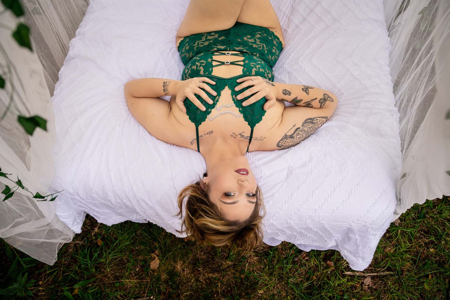 Overhead view of tattooed woman in green lace lingerie lying on white bed in outdoor garden.
