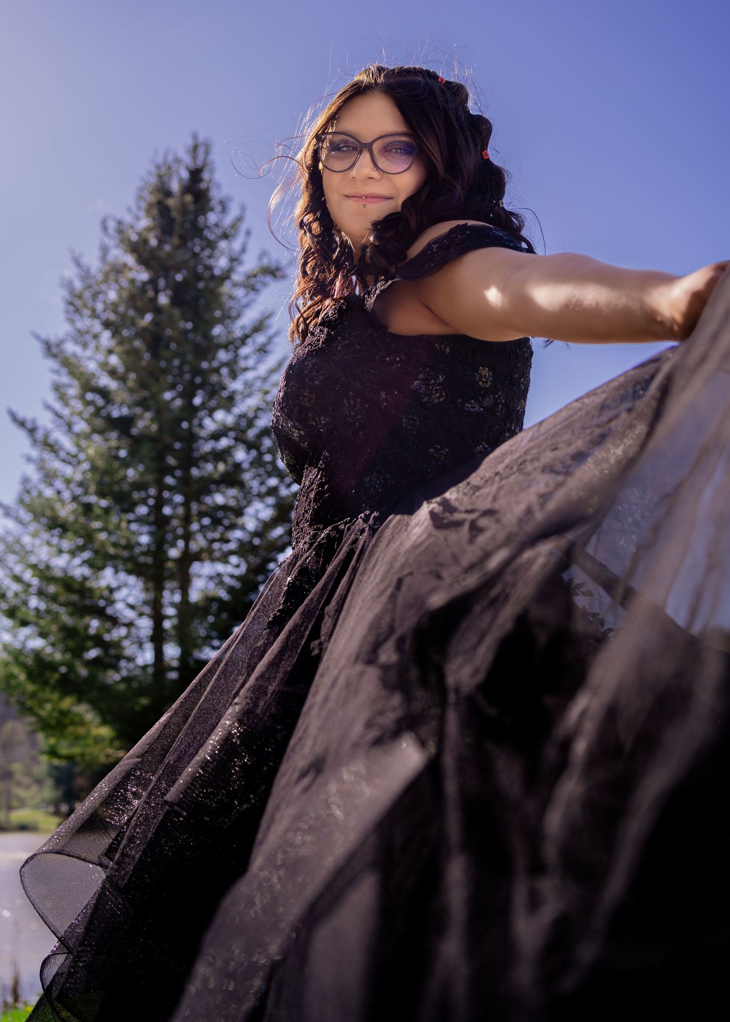 Young woman in a black off-shoulder ball gown twirling outdoors with pine trees in the background.