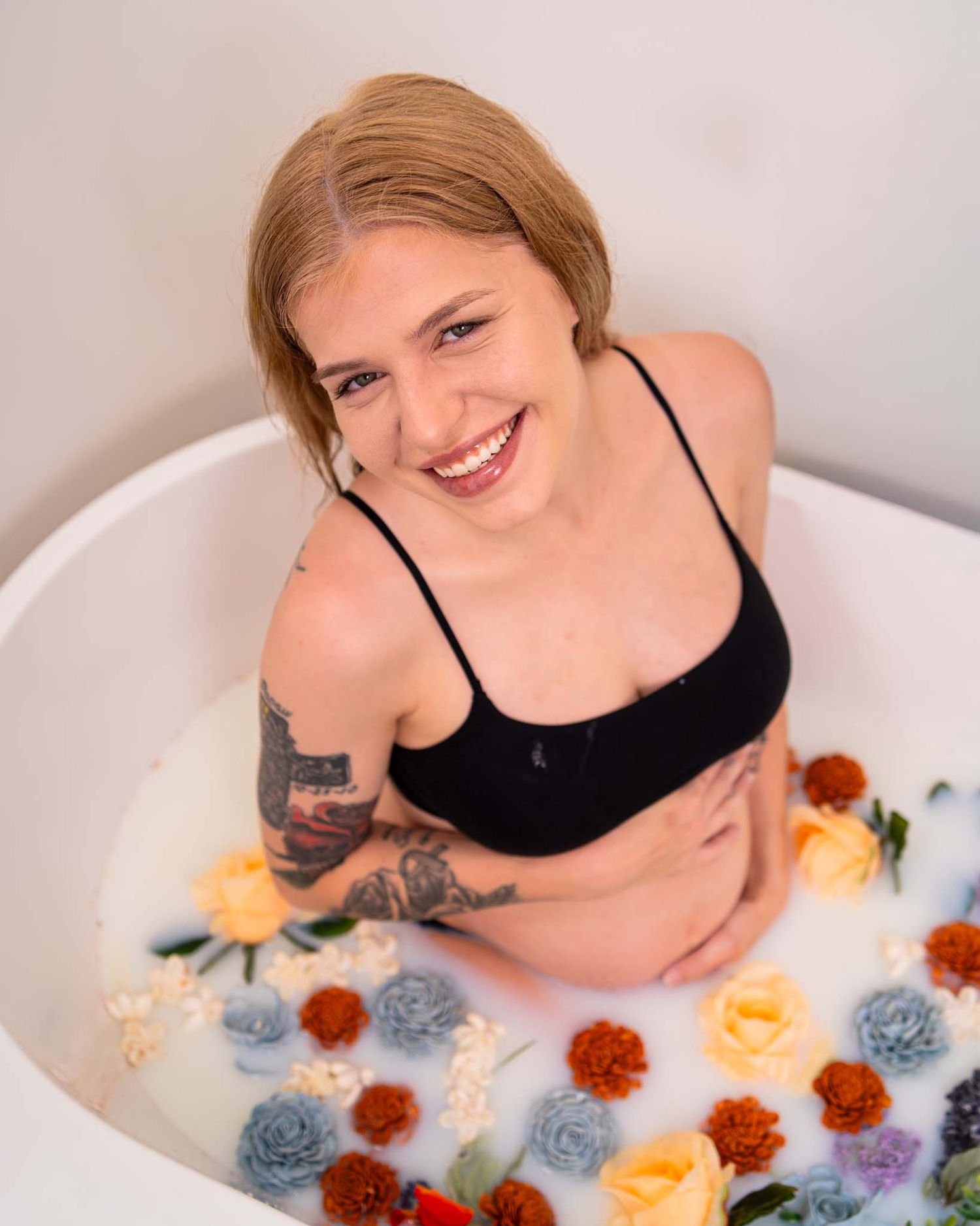 Smiling tattooed pregnant woman in black top sitting in a floral milk bath.