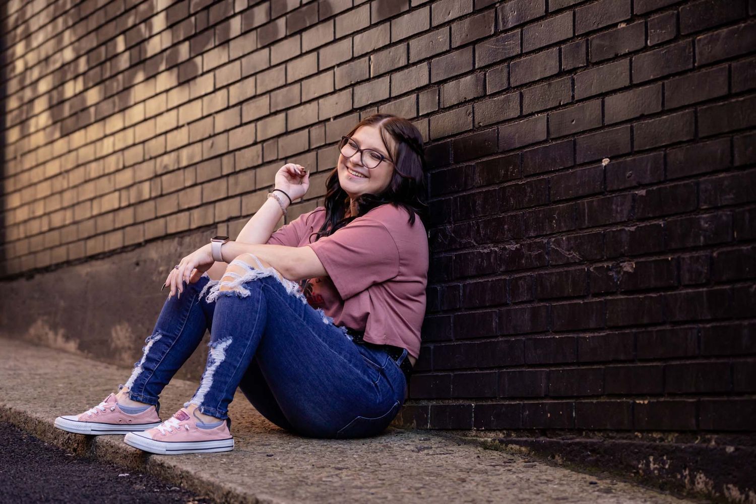 Smiling teen girl in ripped jeans sitting against a brick wall on a sidewalk outdoors.
