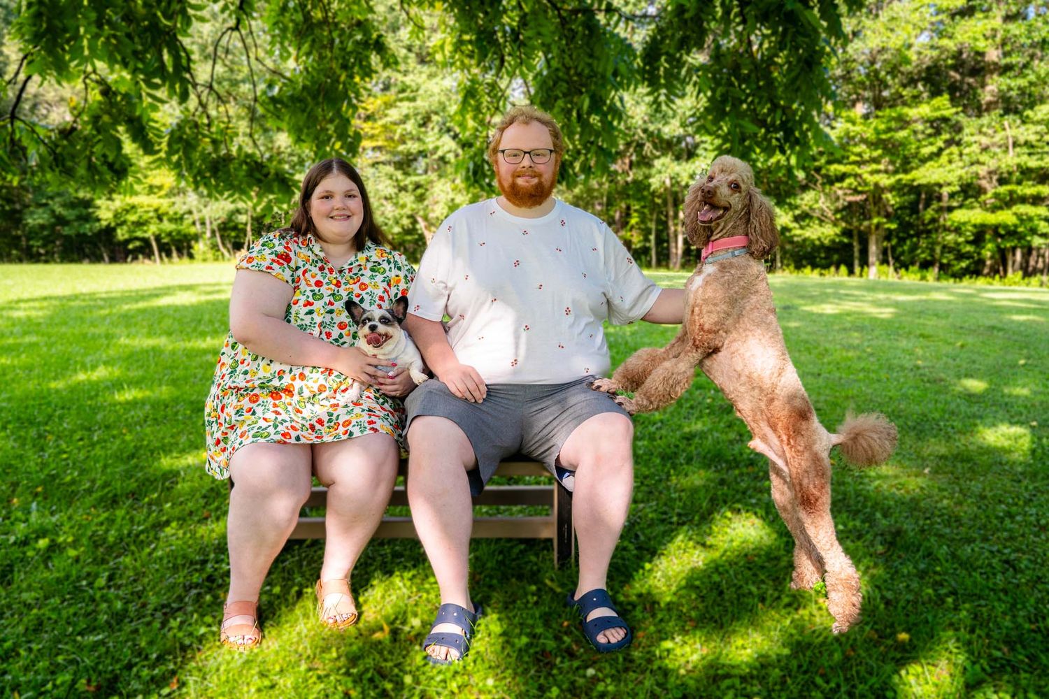 Couple sits on wooden bench in green park with large fluffy dog standing beside them on sunny day.