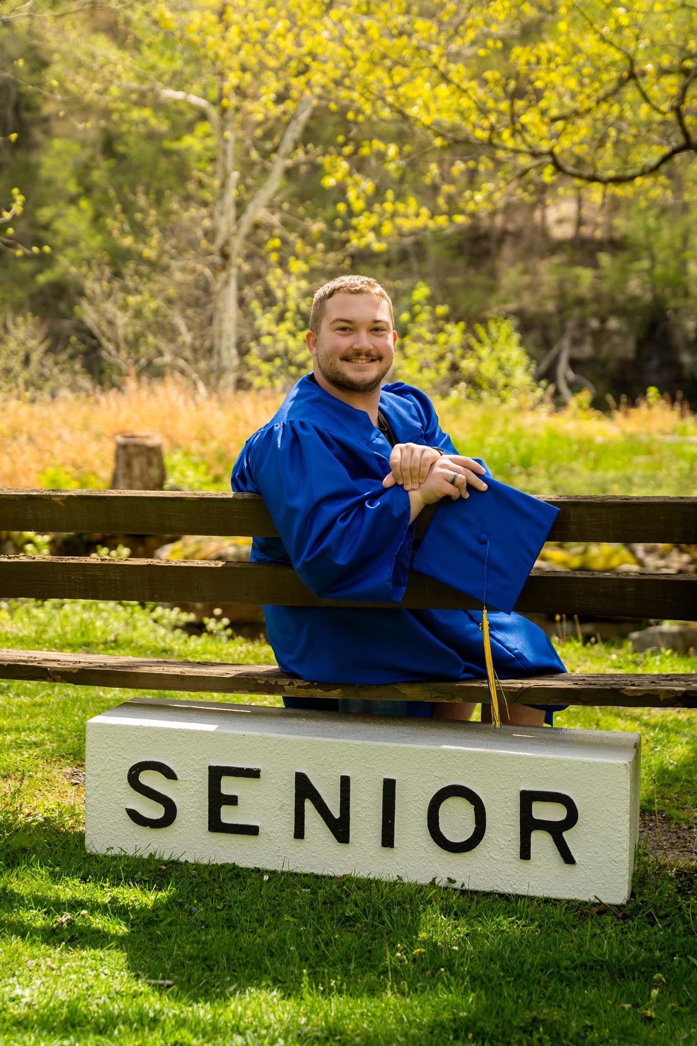 Smiling male senior in blue graduation gown and cap sits on a bench beside a white Senior sign outdoors.