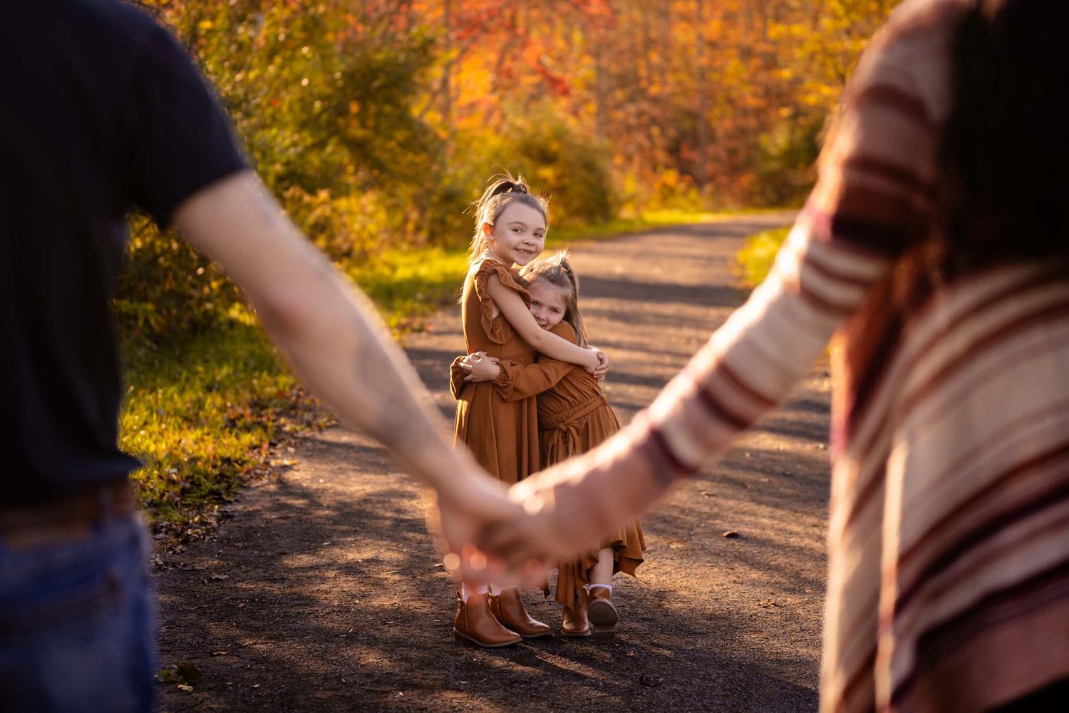 A young girl in a brown dress smiles on an autumn path while her parents hold hands in the foreground.
