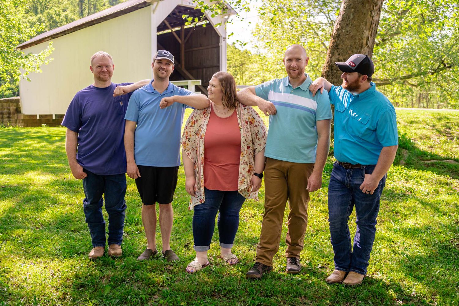 Group of five adults laughing and posing playfully on green grass in front of a covered bridge.