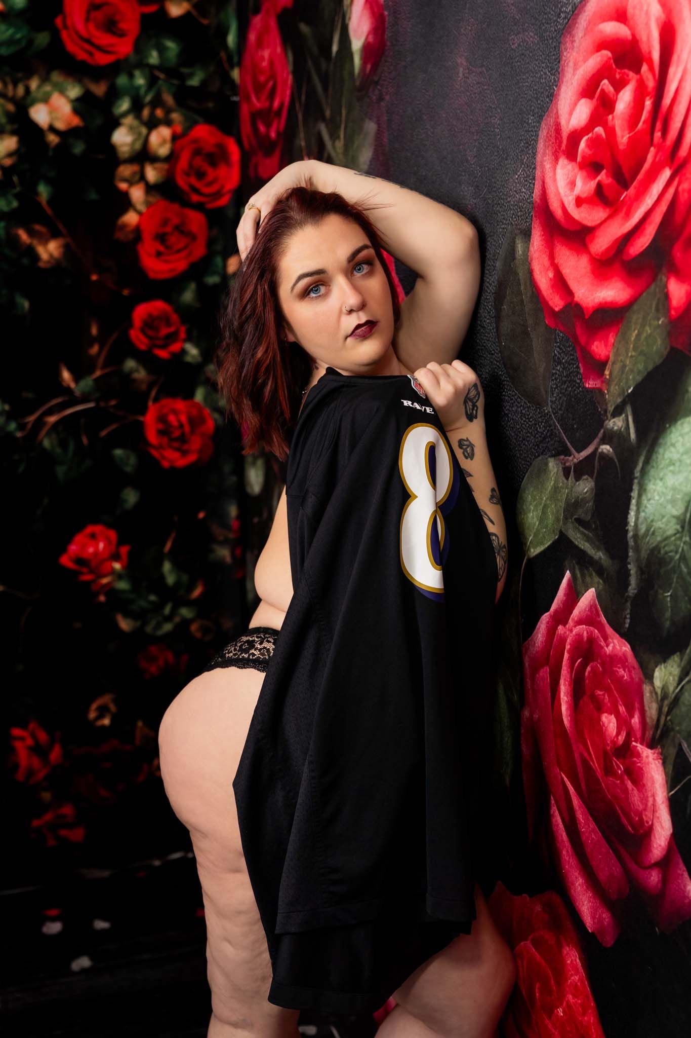 Woman in black NFL jersey poses against vibrant rose backdrop, showing tattoos and lace shorts.