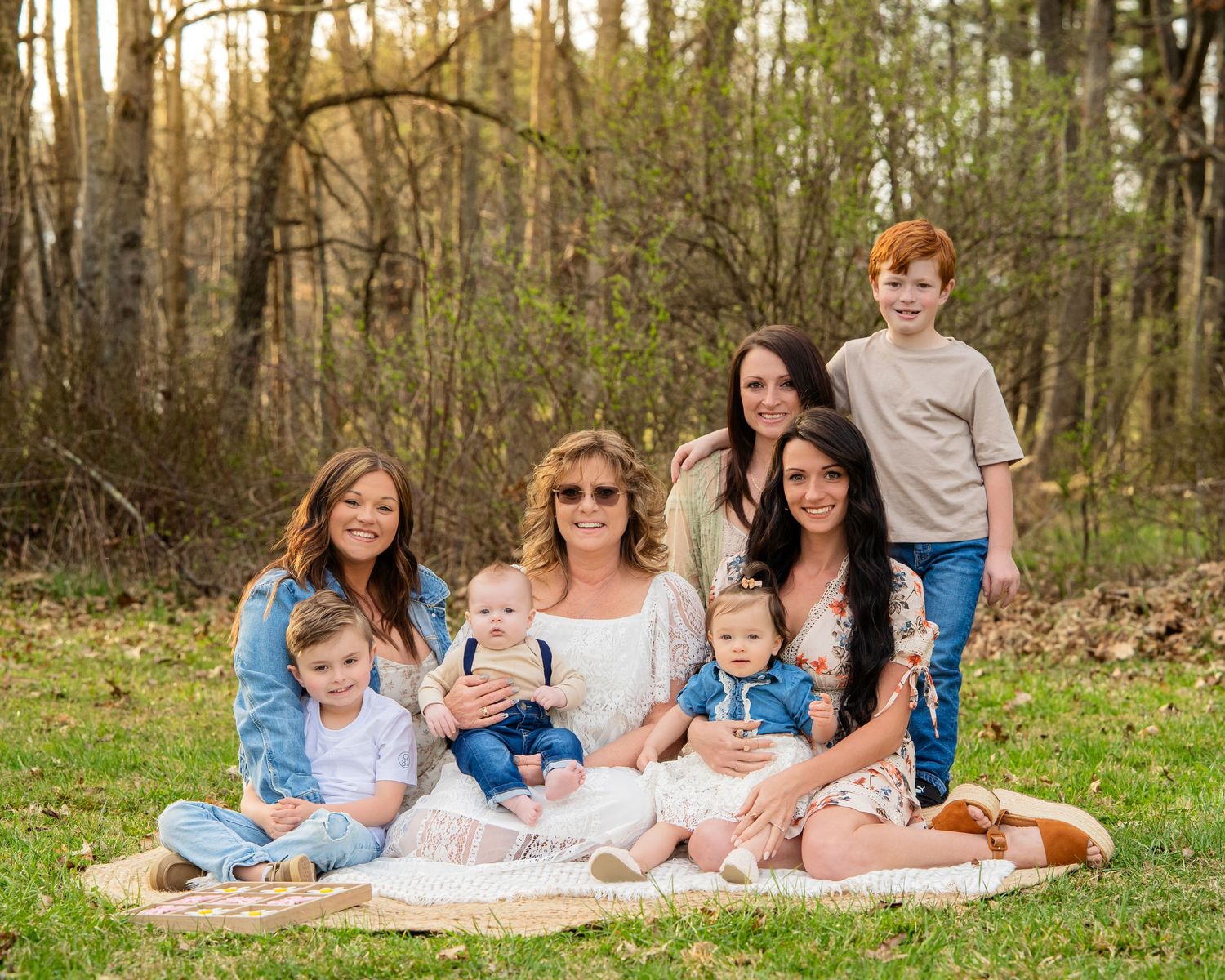 Three generations of family posing together on blanket in wooded outdoor setting.