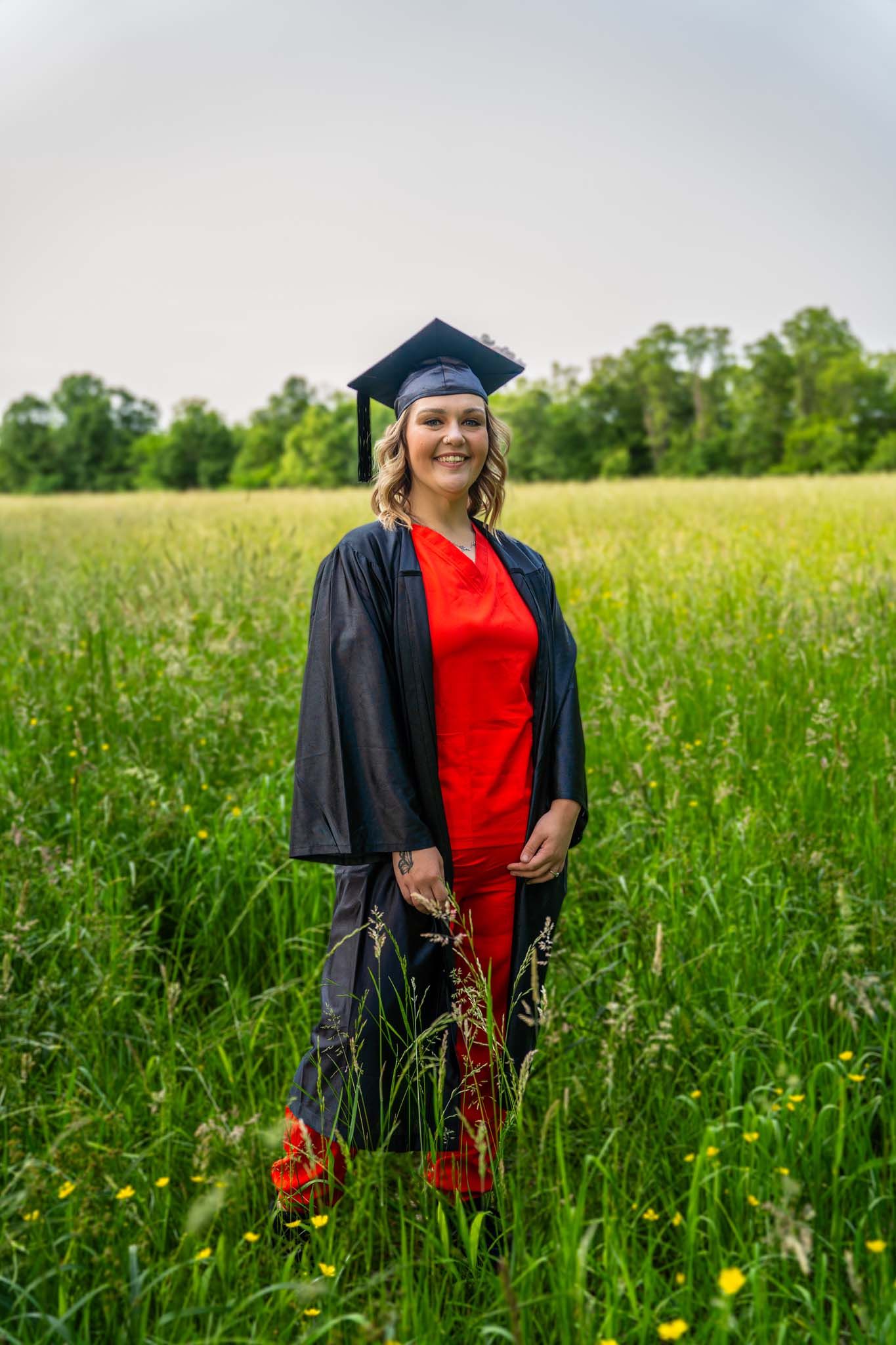 Female graduate in black cap and gown with red outfit standing in green field.