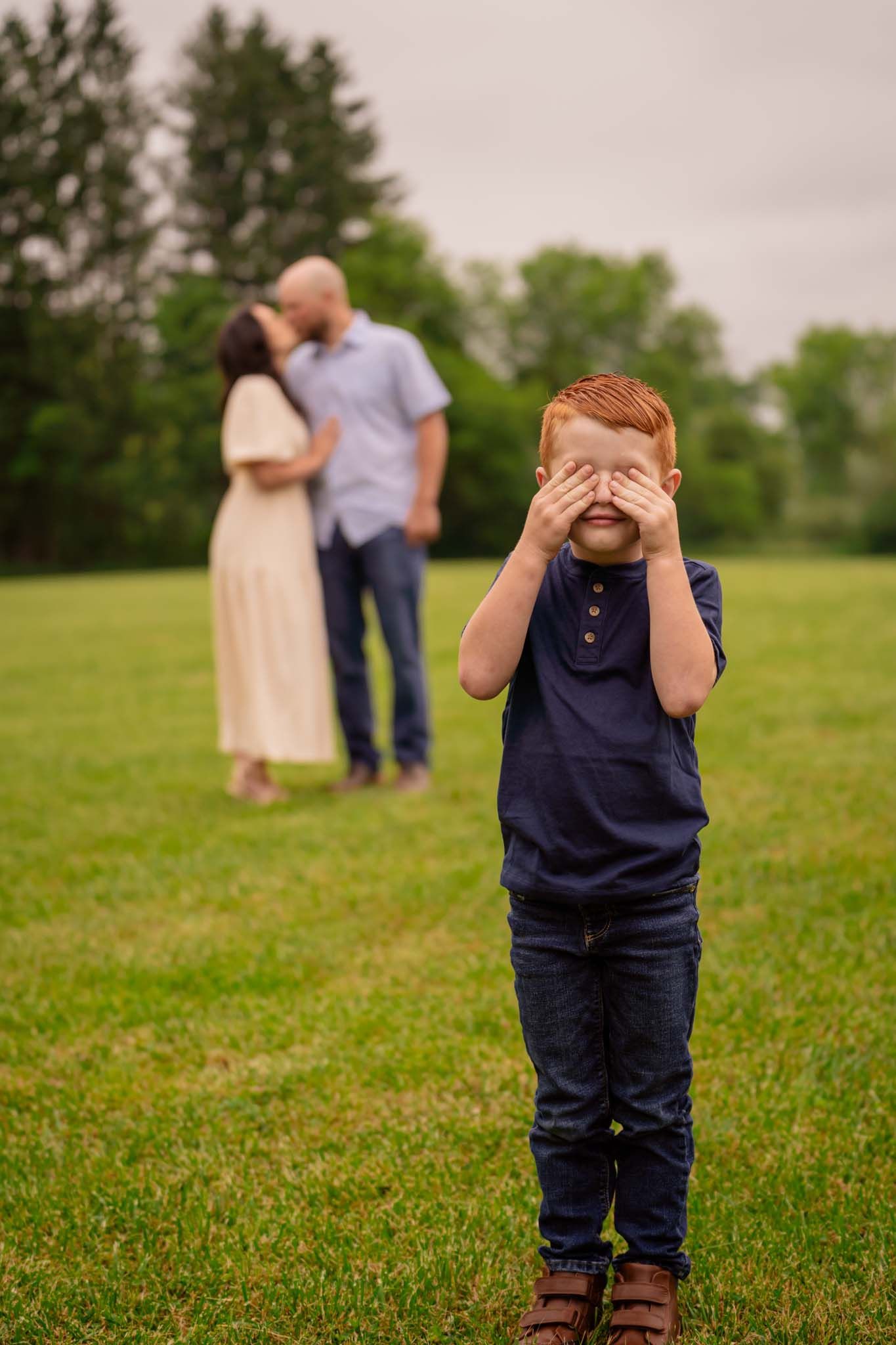 Playful family photo with redhead boy covering eyes in foreground while parents kiss on green lawn behind him.