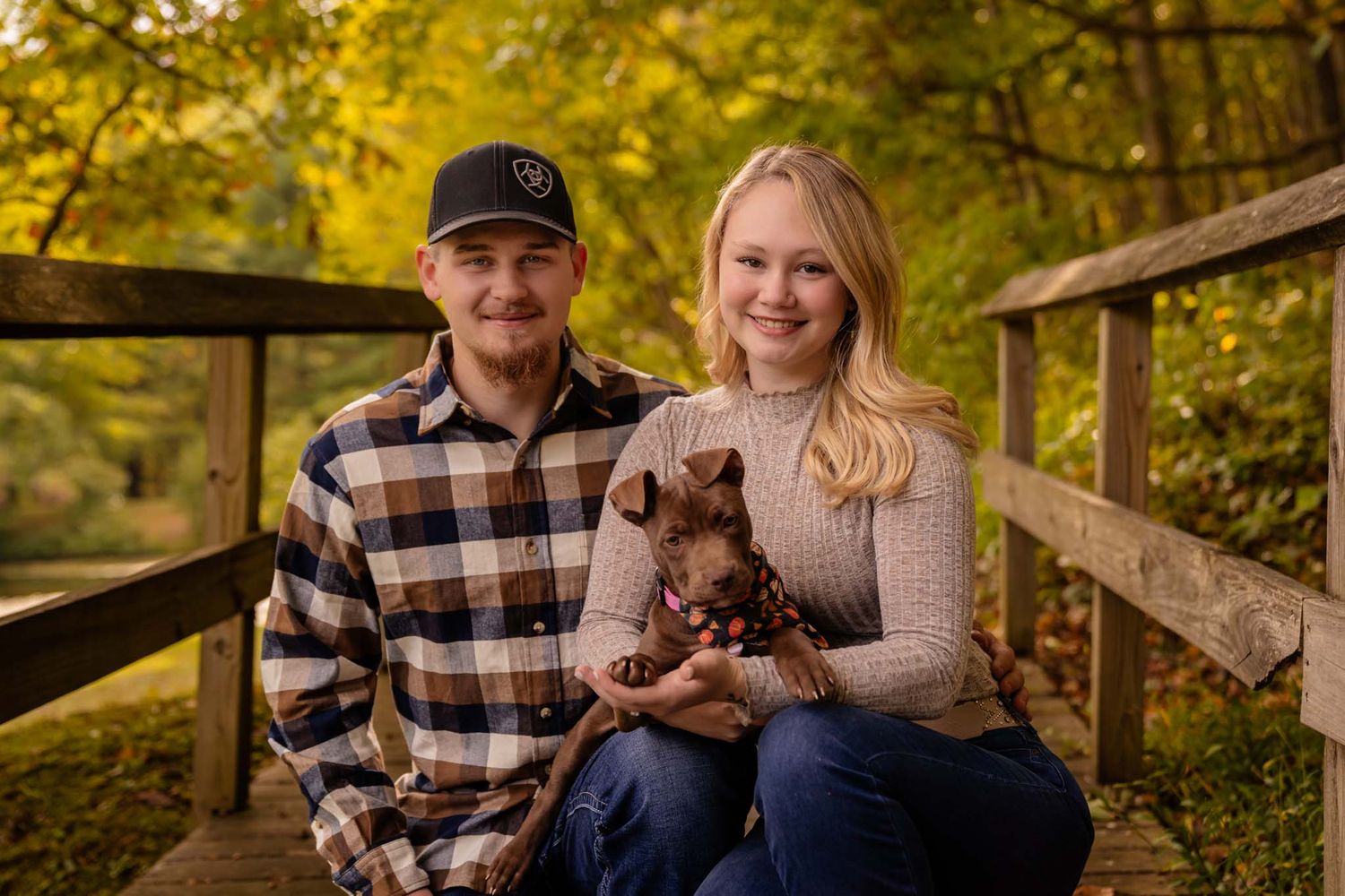 A smiling couple poses on a wooden bridge in autumn, holding their small brown puppy wearing a festive bandana.