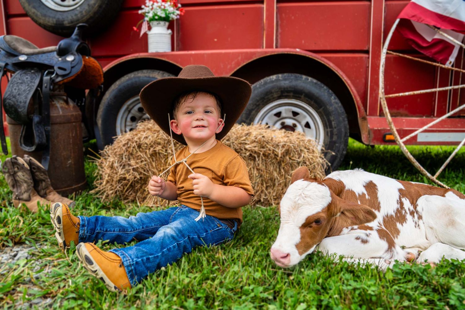 Young boy in cowboy hat sits with baby calf on hay bale near red barn vehicle.