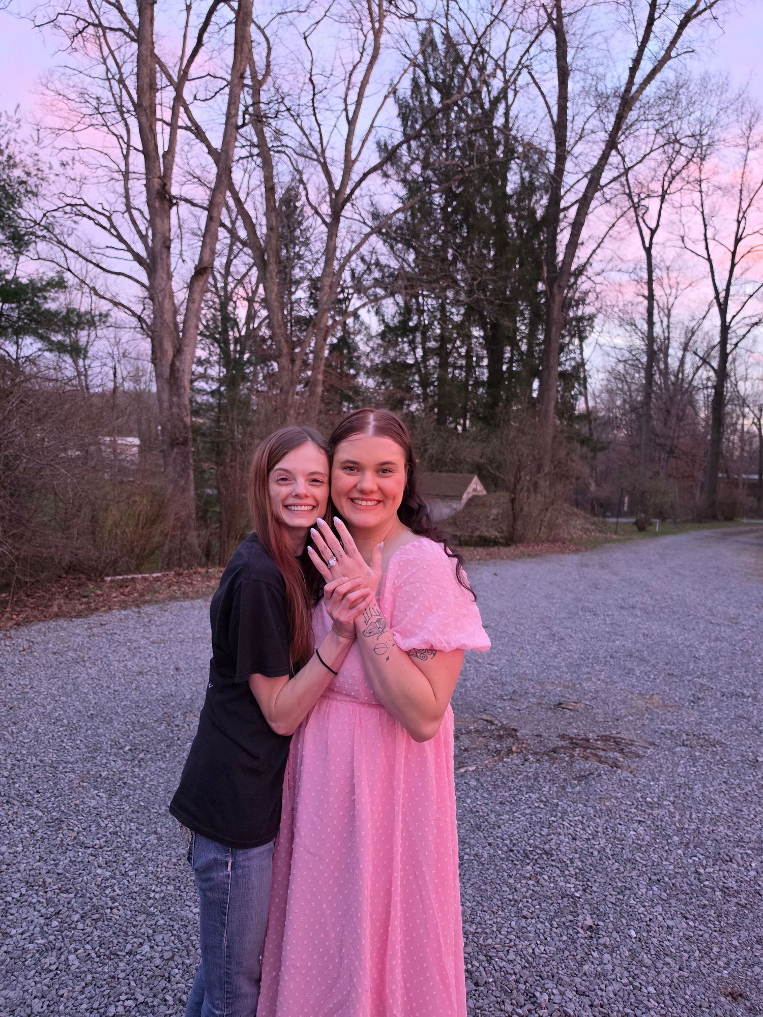 Two young women smiling outdoors at sunset, one in pink dress showing off a ring, trees and gravel road behind them.