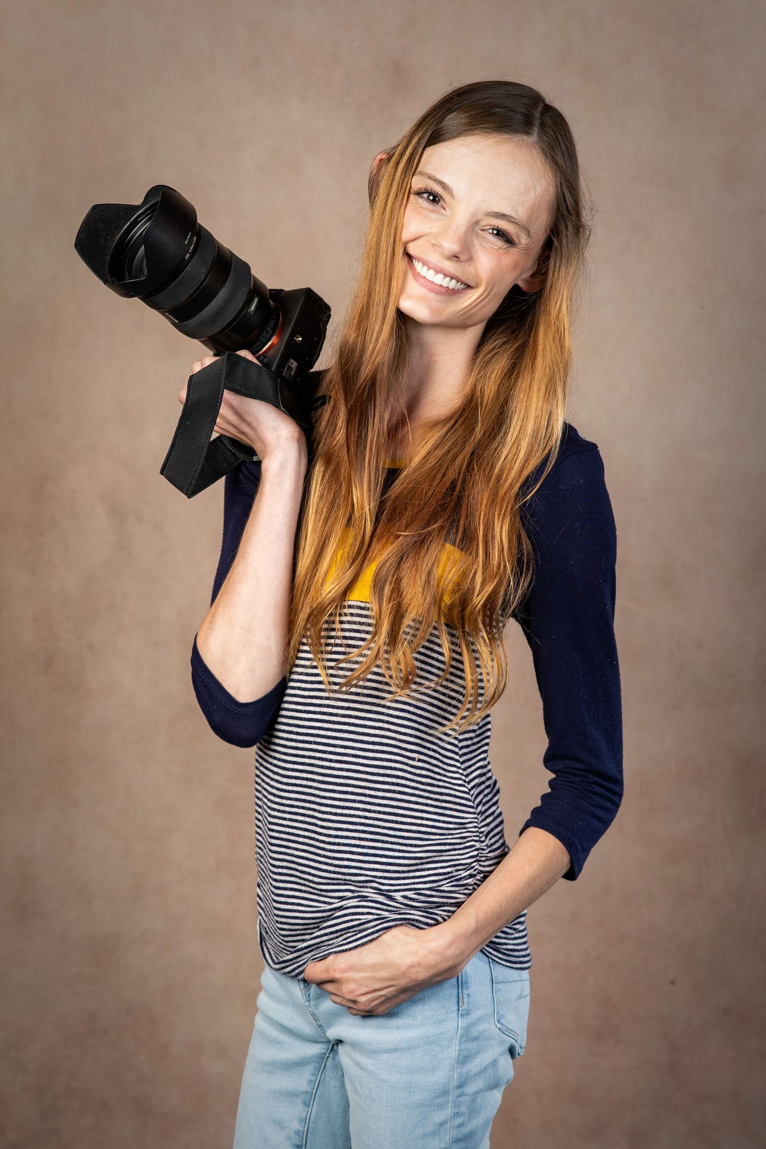 Smiling young woman with long red hair holding a DSLR camera, wearing a striped shirt and jeans in a studio.