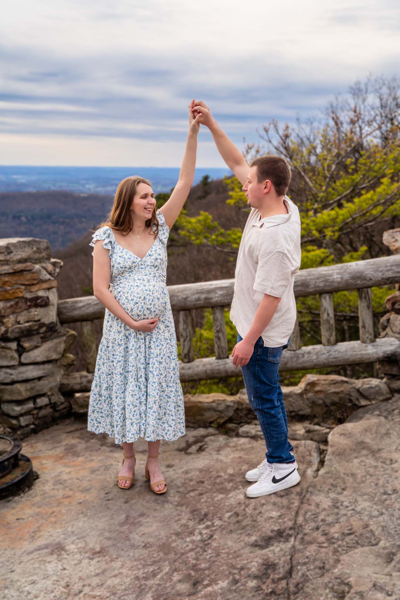 Happy expectant couple dances on rocky overlook with scenic mountain valley backdrop.