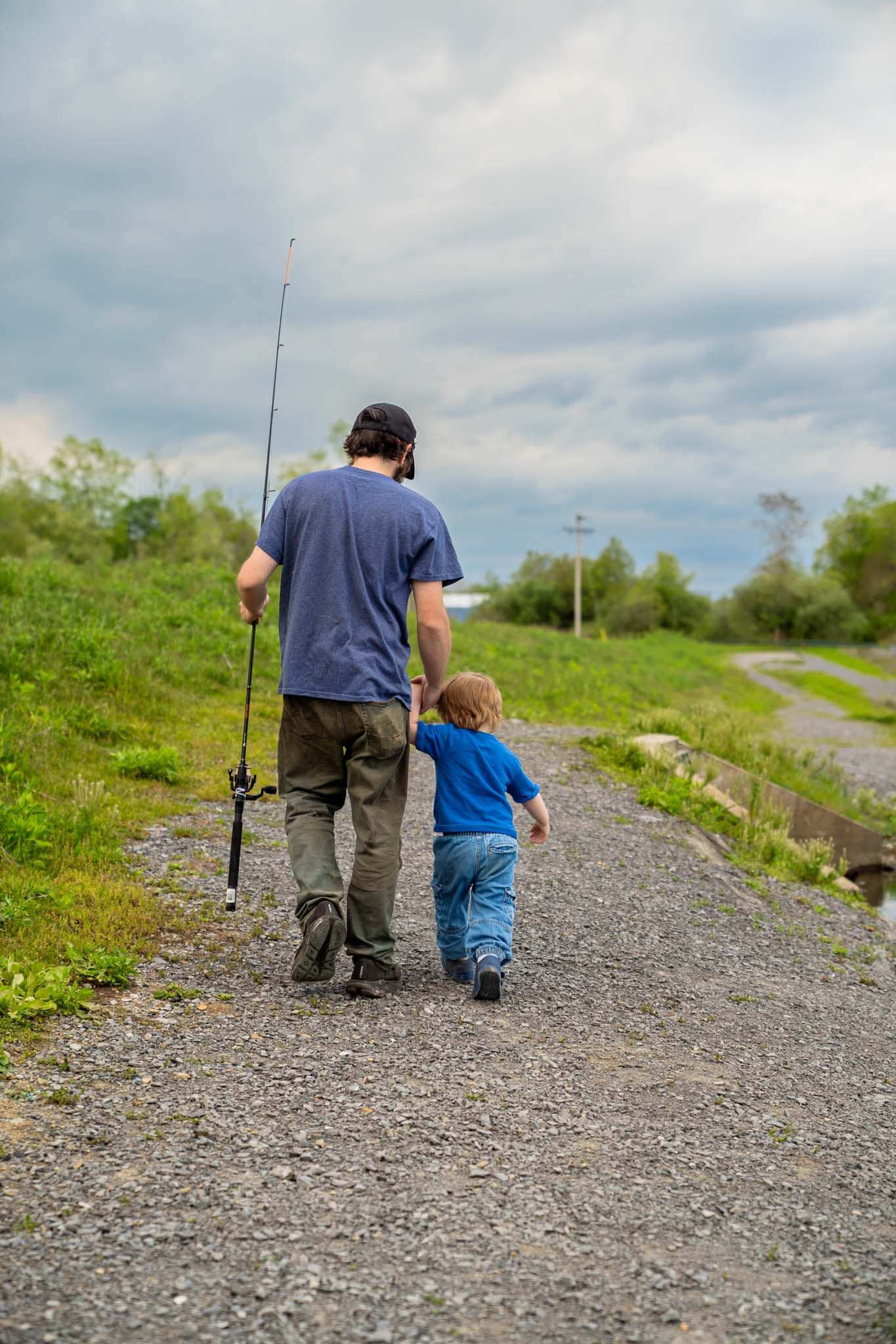 Father and young child walking hand in hand on gravel path carrying fishing rod under cloudy sky.