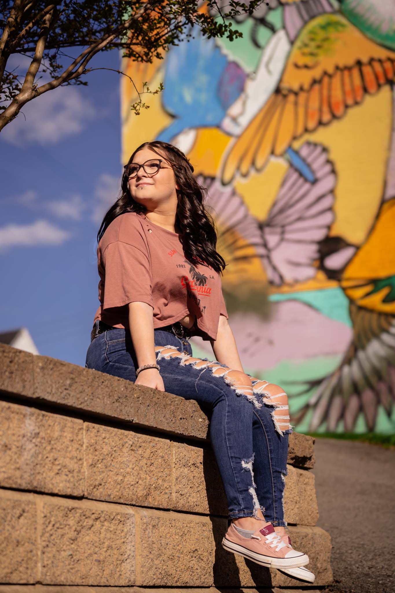 Young woman in ripped jeans and pink top sitting on stone steps before a colorful bird mural.