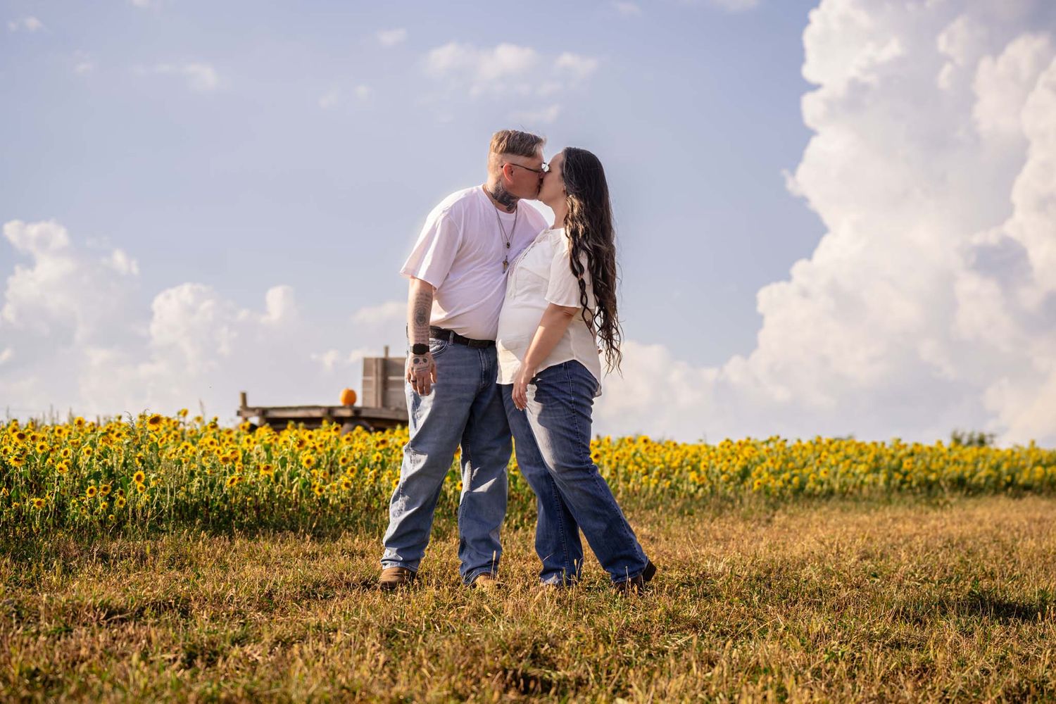 Couple sharing a kiss in a sunflower field with dramatic clouds and farm equipment in background.