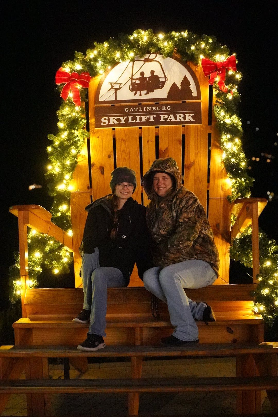 Couple sitting in giant wooden chair at Gatlinburg Skylift Park, decorated with Christmas lights and red bows at night.