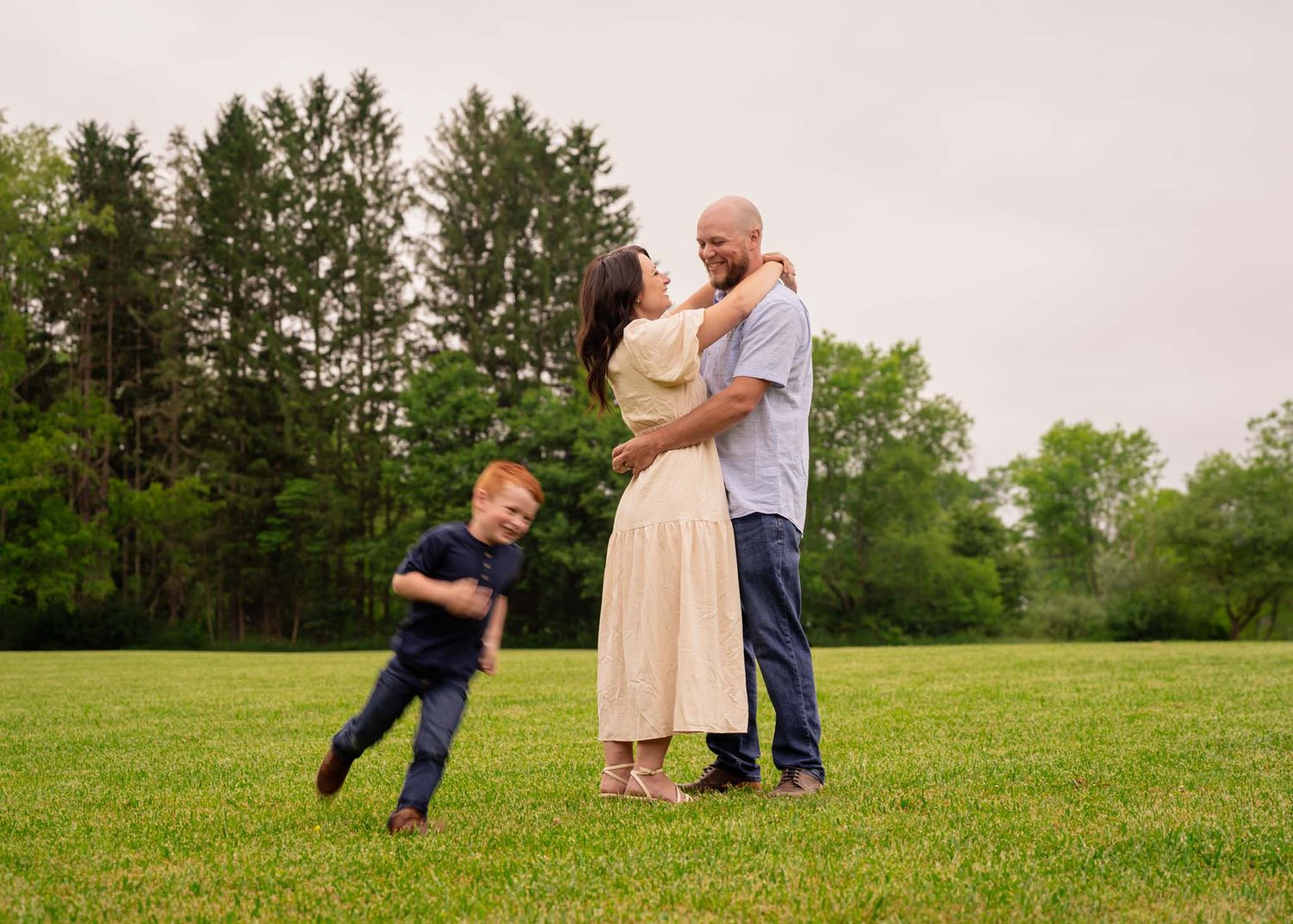 A happy family poses outdoors on green grass with trees behind them as a young redheaded boy plays nearby.