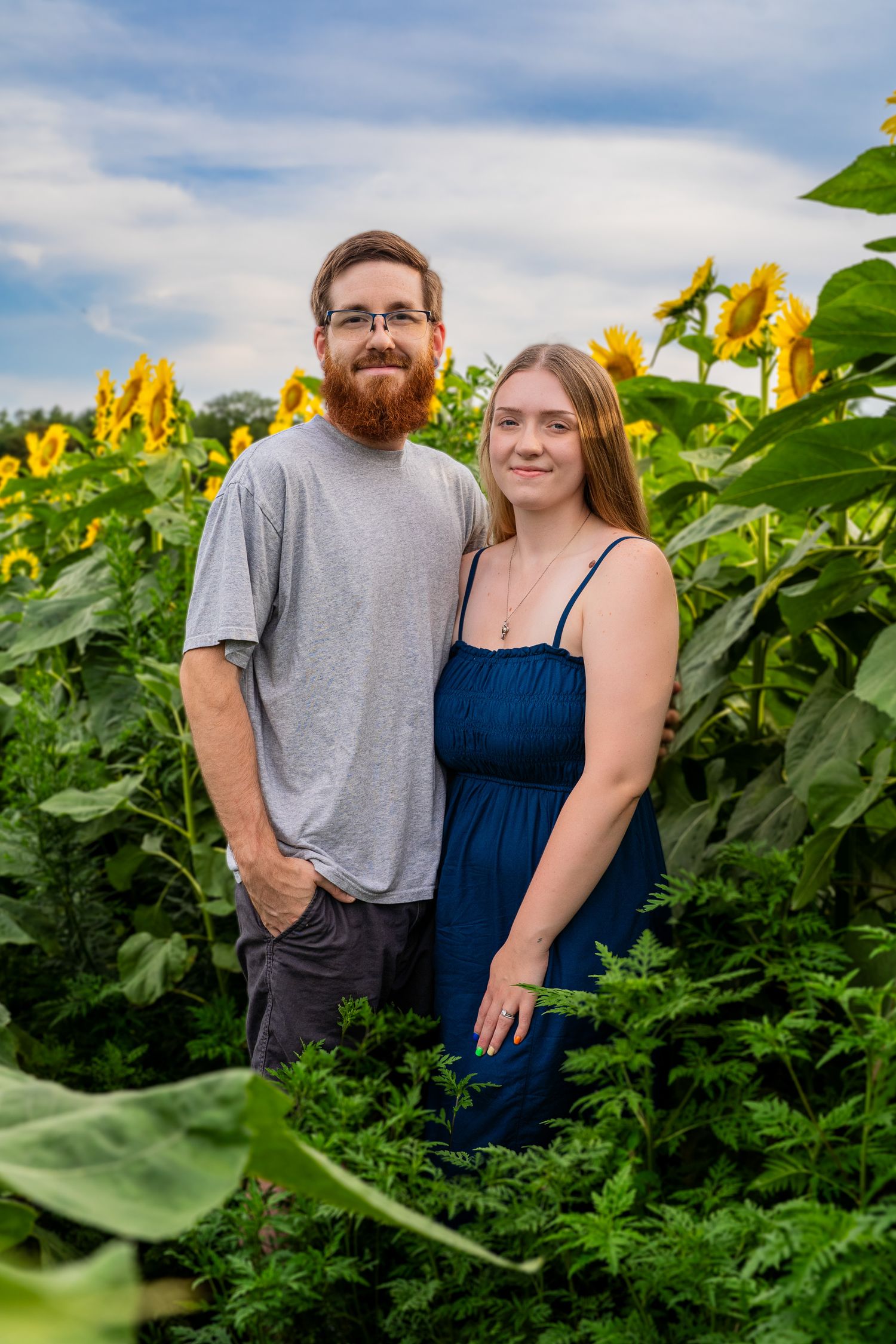 Smiling couple poses together in a vibrant sunflower field under a blue sky.