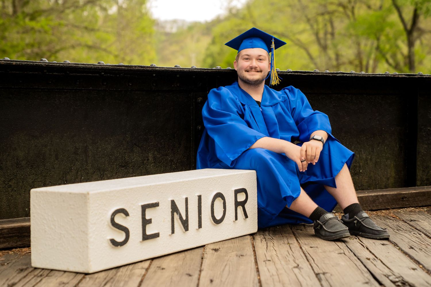 Happy graduate in blue cap and gown sits beside a Senior sign on wooden bridge.