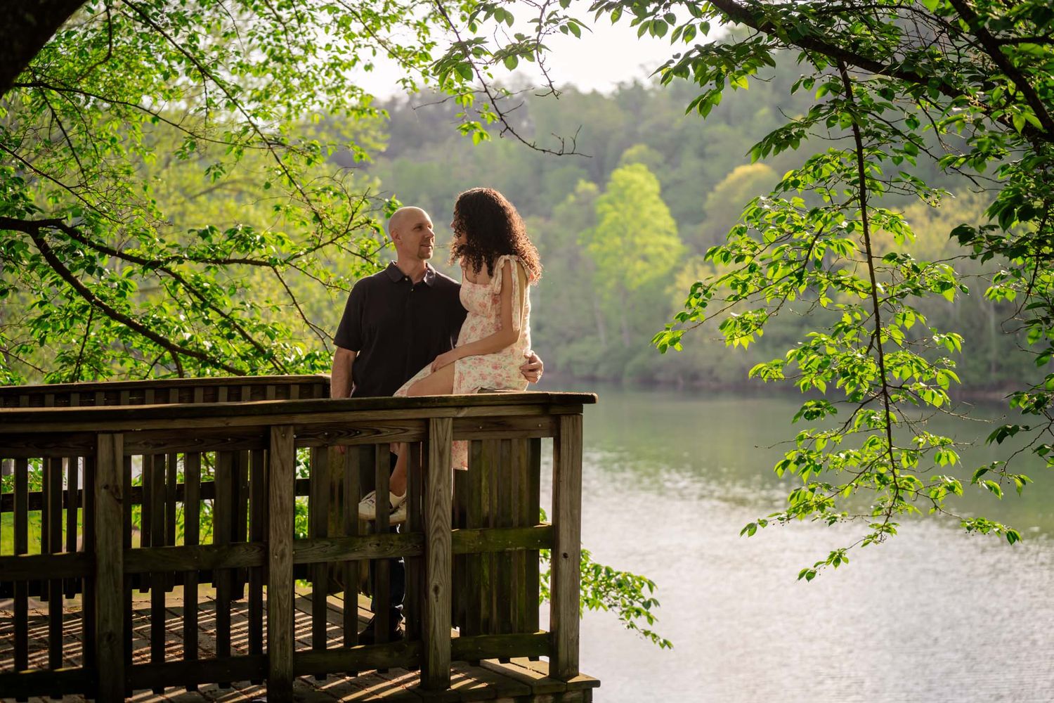 Couple standing on a wooden deck overlooking a serene lake surrounded by lush green trees.
