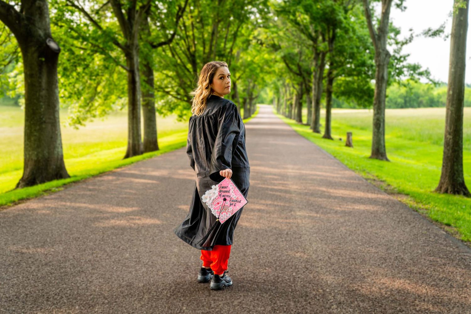 Graduate in black gown holding pink cap walking down tree-lined path.