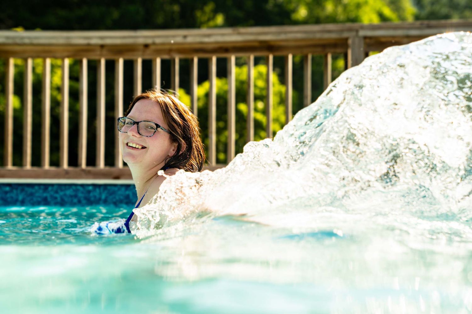 Young woman with glasses smiles joyfully beside pool water feature on bright sunny day.