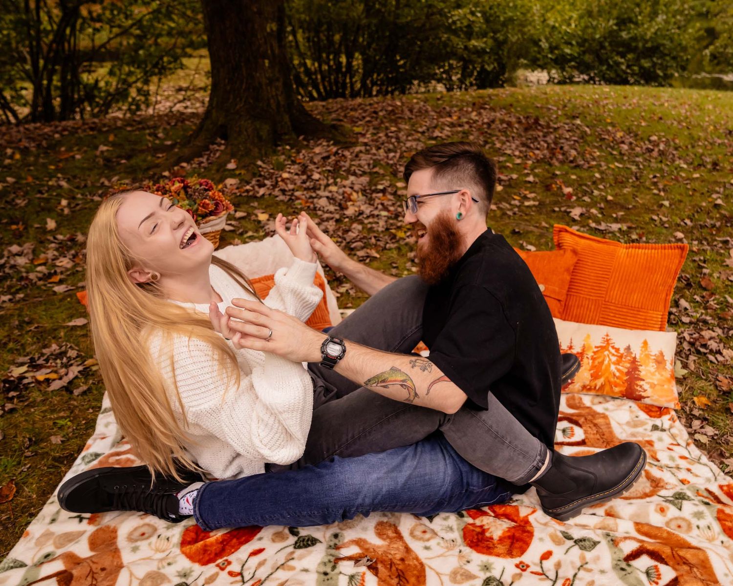 Couple laughing together on a floral blanket in an autumn park setting with orange pillows and fallen leaves.
