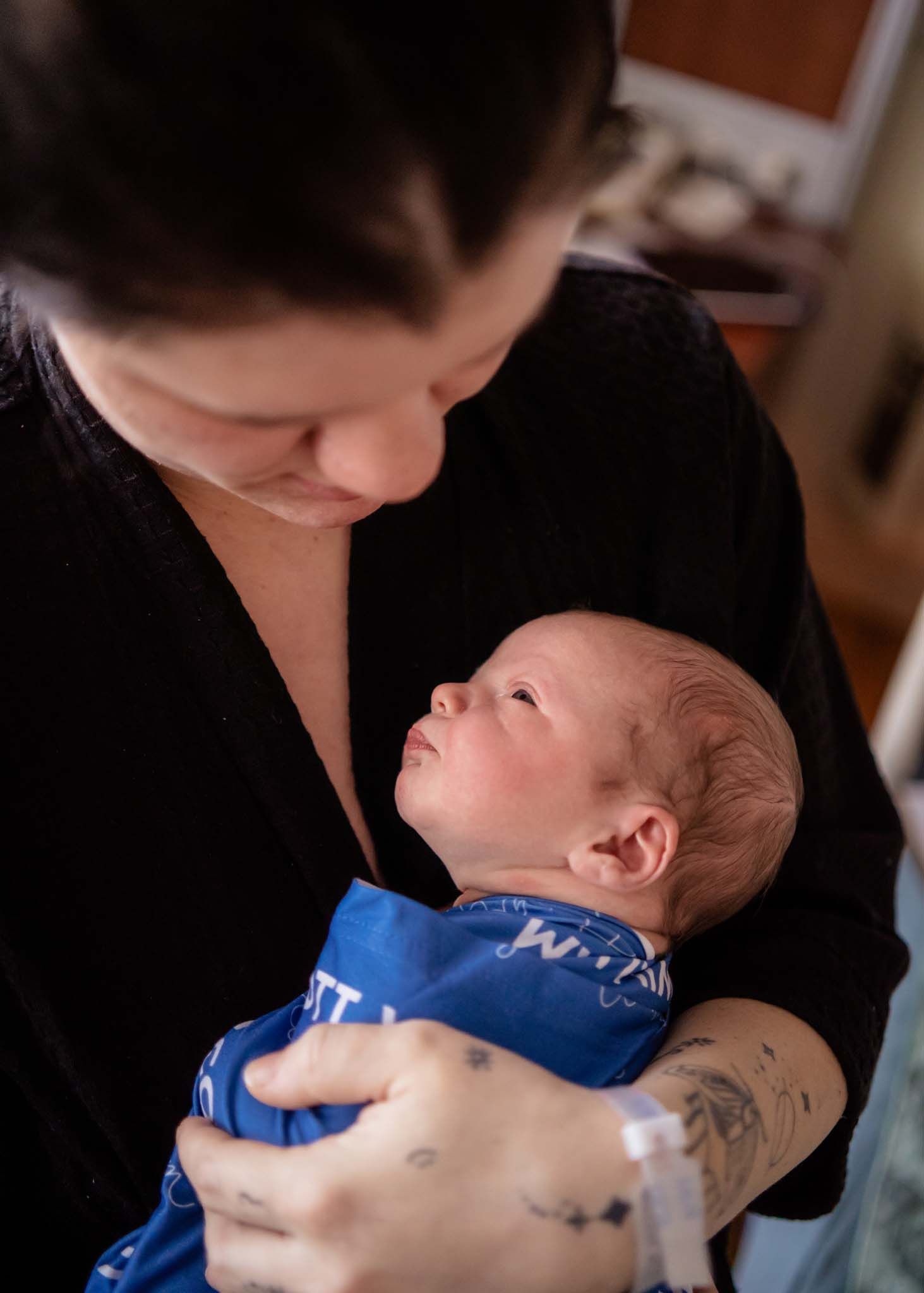 A tattooed mother tenderly holds her newborn baby wrapped in a blue blanket in a hospital room.