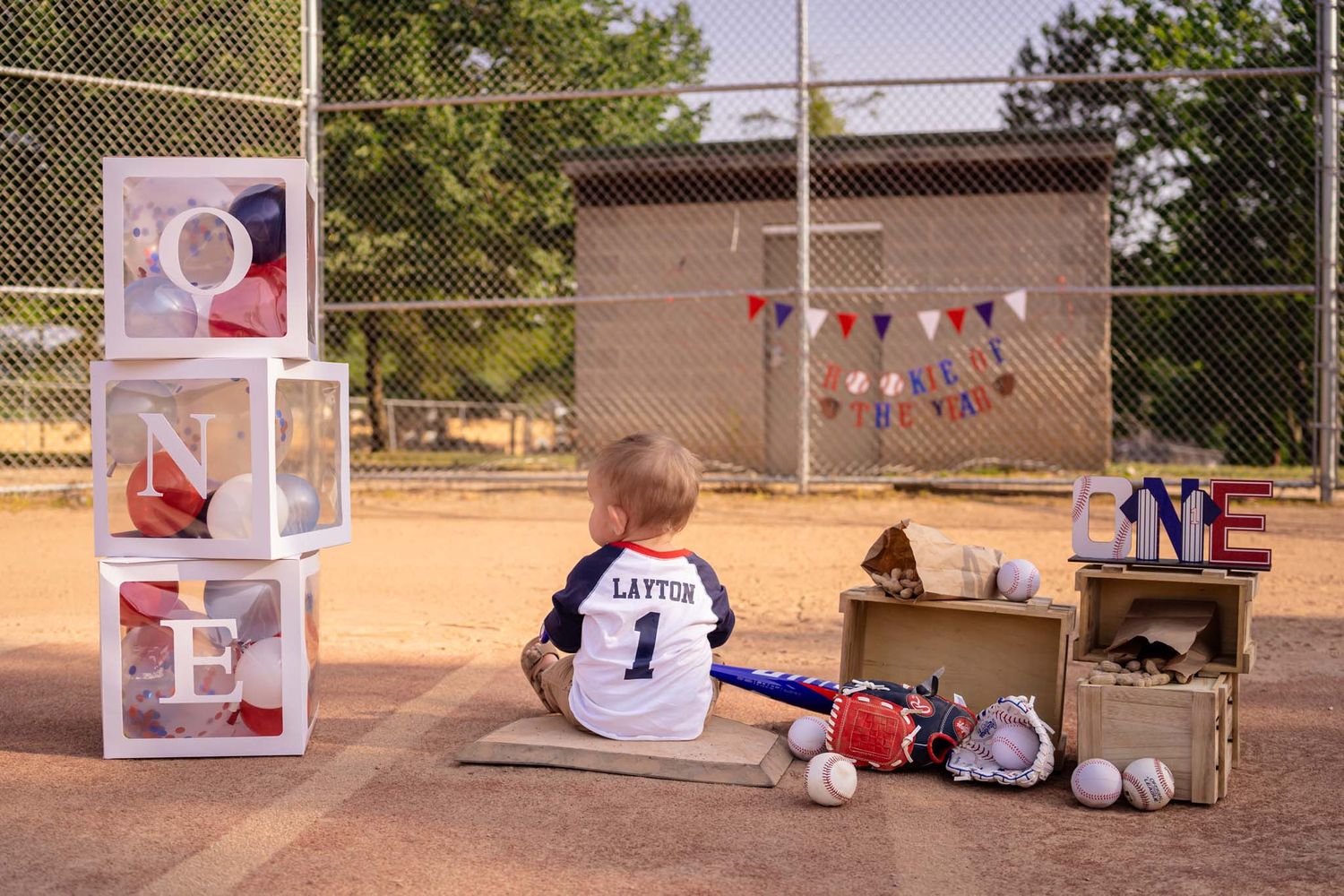 Toddler in baseball jersey sits on diamond with ONE blocks, bat, glove, and bunting for a first birthday photo.