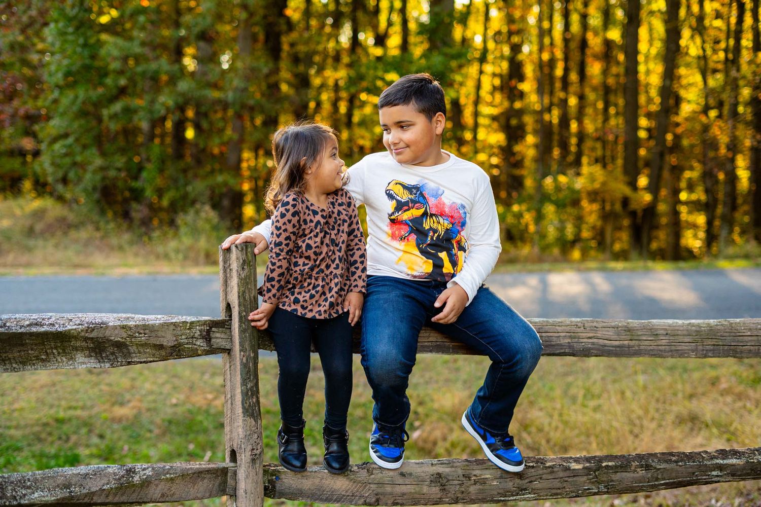 Two children sit on a rustic wooden fence surrounded by autumn foliage, smiling at each other outdoors.