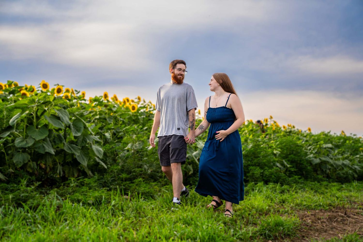 Couple walking through sunflower field, woman in navy dress holding flowers, man in gray shirt, under cloudy sky.