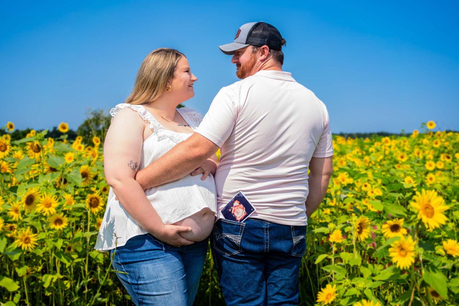 A couple stands back to back in a sunflower field with an ultrasound photo in his pocket.